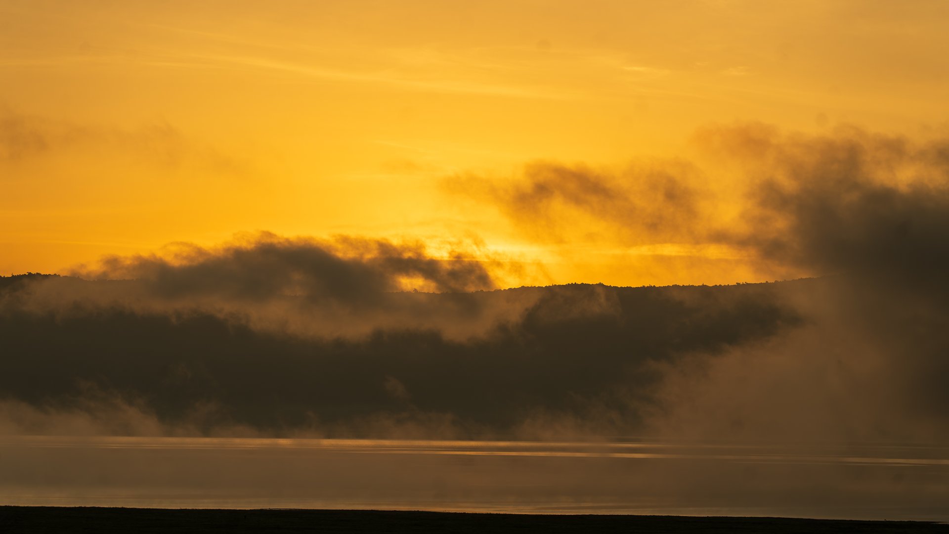 Golden sunrise in Ngorongoro crater over misty mountains and calm water, creating a serene landscape.