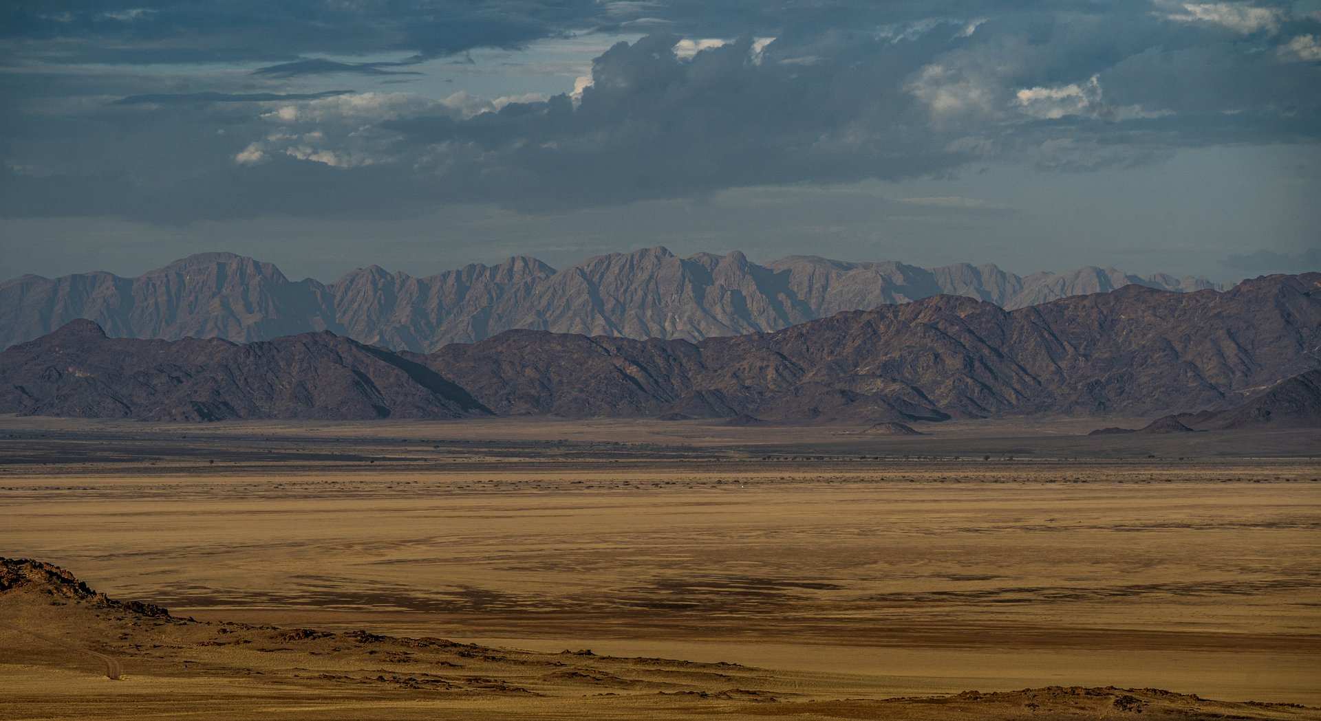 Expansive desert landscape near Sossusvlei, Namibia, with a golden-brown plain stretching toward multiple layers of rugged mountains beneath a dramatic cloud-filled sky.