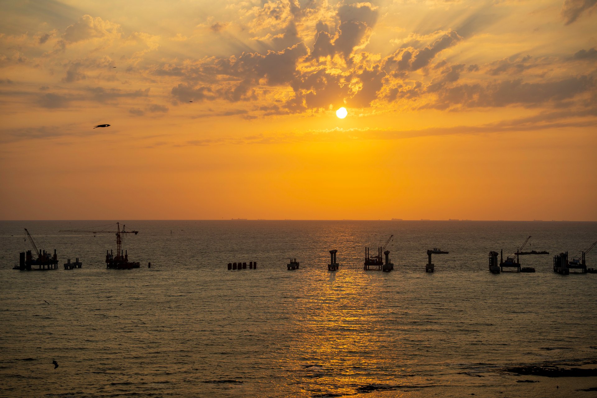 bandra-bandstand-sunset-mumbai-coastal-road-construction Sunset over the ocean with construction platforms and cranes silhouetted against the sky.